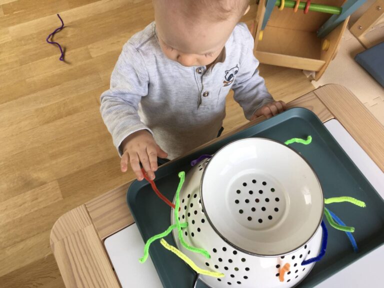 Ein Baby zieht Pfeifenputzer aus den Löchern eines Nudelsiebs